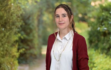 Portrait of a young woman with long dark hair, wearing a white blouse and red jacket, in front of a green wooded background