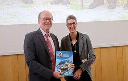 In the lecture hall: A man and a woman stand next to each other and smile into the camera. The man is holding a large map.