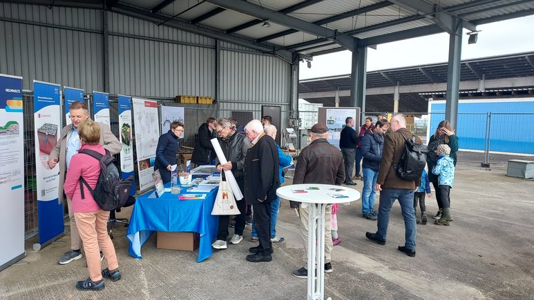 Unter einem halboffenen Hallendach sind Tische mit Infomaterial, Roll-ups und Poster aufgebaut. Davor stehen interessierte Besucher jeden Alters im Dialog mit den Forschenden.