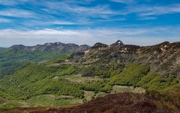 in the front green meadows and forests, in the background rugged brown vulcano chain, blue sky with cirrostratus clouds