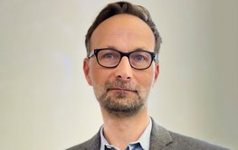 shirt, grey patterned jacket and black glasses in front of grey wall