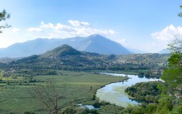 A green landscape. Middle and background: mountains. A river meanders in the foreground.