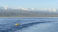 The picture shows a buoy with a GNSS antenna in the centre, which is being pulled across the lake behind a ship with a rope. The snow-covered mountains of Tian Shan can be seen in the background.