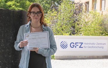 The picture shows Candice Filoche holding the DMG award in her hands. In the background, you can see a column from the Column Forum and the GFZ logo at the entrance to House G.