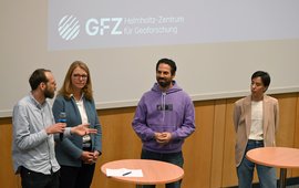 Four people are standing at a table in front of a screen and discussing.