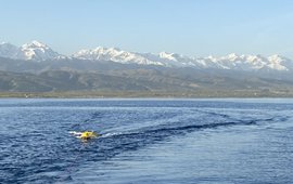 The picture shows a buoy with a GNSS antenna in the centre, which is being pulled across the lake behind a ship with a rope. The snow-covered mountains of Tian Shan can be seen in the background.