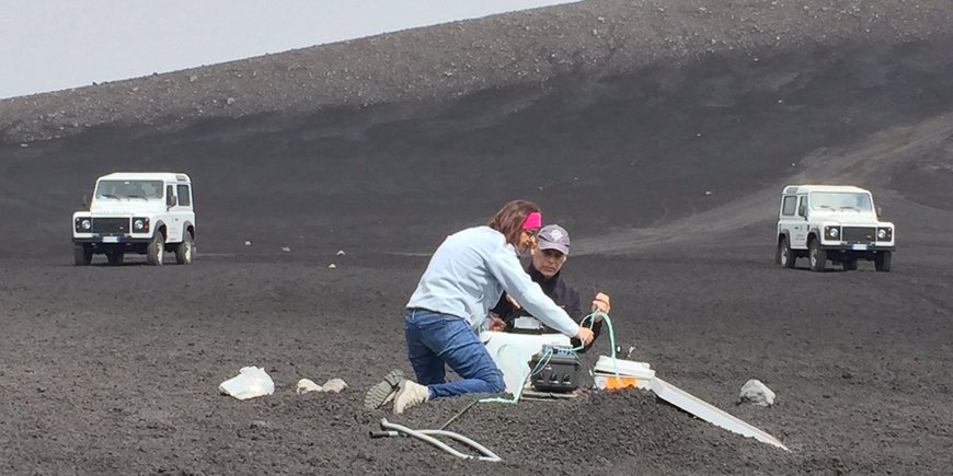 Two people kneel in the grey cinders of the volcano and connect two fibre optic cables.