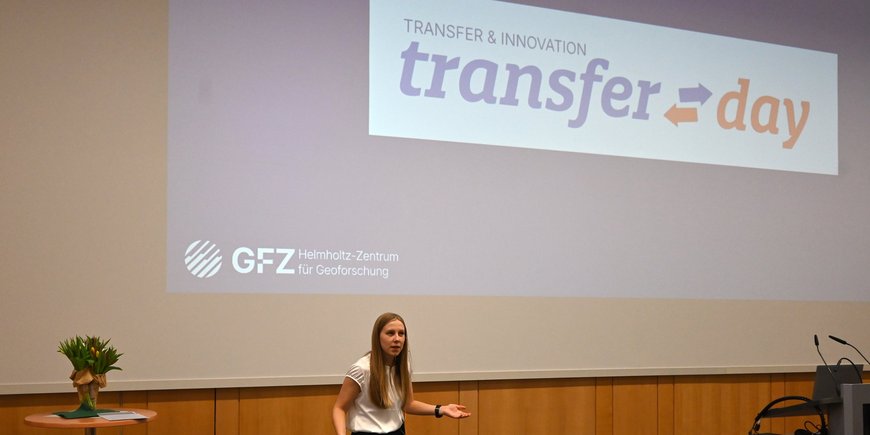 Eine Frau steht vor einer Leinwand und moderiert eine Veranstaltung. Auf die Leinwand ist "Transfer Day" projiziert.