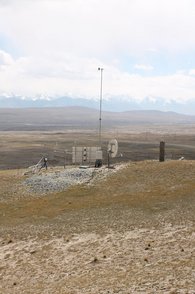 The picture shows a view over the landscape of the Tian Shan. In the centre is a measuring station with a GNSS station (right), a wind mast (centre), in front of it the box with instruments and on the left a snow measuring system.