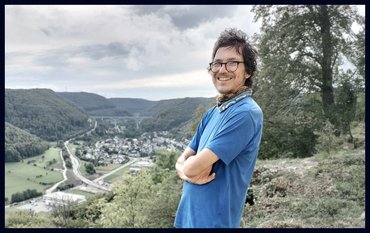 A young man with dark hair and glasses stands on a green slope. A town in the valley.