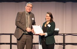 A middle-aged man wearing a brown suit hands a certificate to a young woman wearing a green jacket and black trousers. Both are standing on a black stage. A grey curtain in the background.