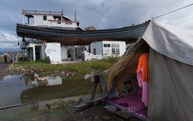 A boat is on the roof of a destroyed house, a tent on the ground, a puddle, clothes hanging on a line, two men standing next to it.