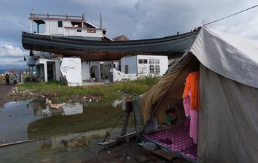 Ein Boot ist auf dem Dach eines zerstörten Hauses, am Boden ein Zelt, eine Pfütze, Kleider hängen an einer Leine, zwei Männer stehen daneben.