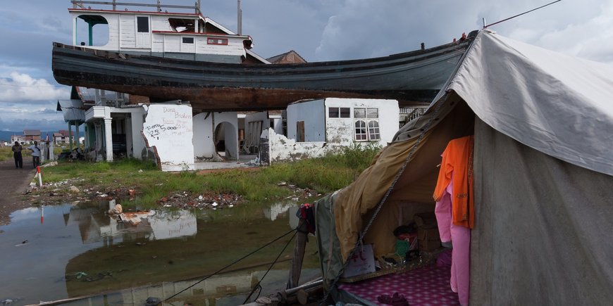 A boat is on the roof of a destroyed house, a tent on the ground, a puddle, clothes hanging on a line, two men standing next to it.