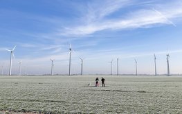 A few wind turbines on flat land. Two people are standing in front of them on the frosted field. Blue sky with hazy clouds.