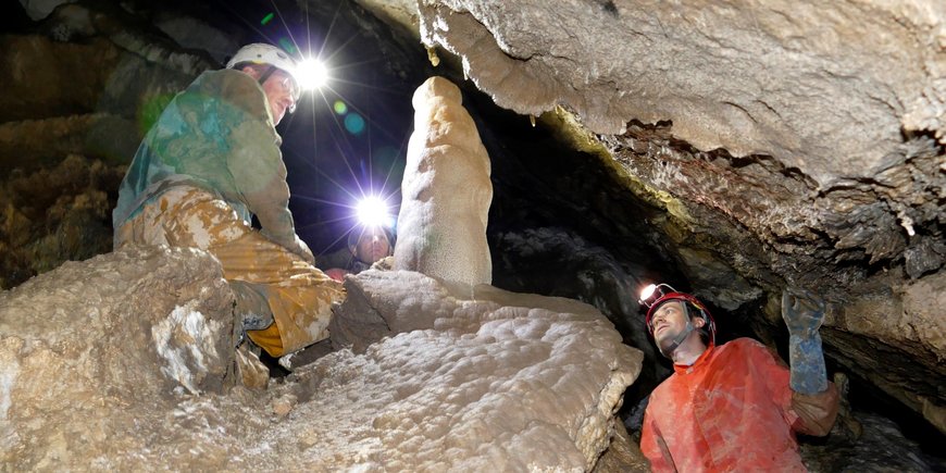 Two people kneel and stand with helmet and headlamp in a narrow stalactite cave