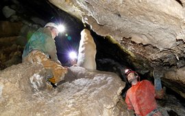 Zwei Menschen knien und stehen mit Helm und Stirnlampe in einer engen Tropfsteinhöhle