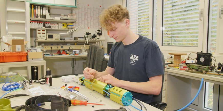 Ein junger Mann sitzt an einem Labortisch mit Kabeln, anderem Material und diversen Werkzeugen. Er schraubt an einer rohrförmigen Apparatur.