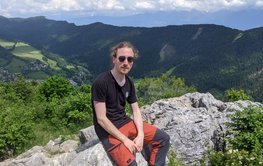 A young man sitting on rocks, green mountains and sky in the background