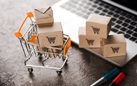 Ein kleiner Spielzeug-Einkaufswagen steht auf einem Laptop, beladen mit kleinen Kisten.