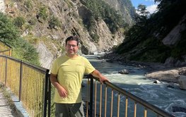 A young man with dark hair and glasses, wearing a yellow T-shirt, stands by a railing. Behind him is a river in the mountains.