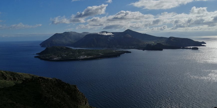 A dark grey volcanic island in the Mediterranean; blue sky with clouds.