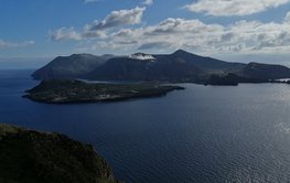 A dark grey volcanic island in the Mediterranean; blue sky with clouds.