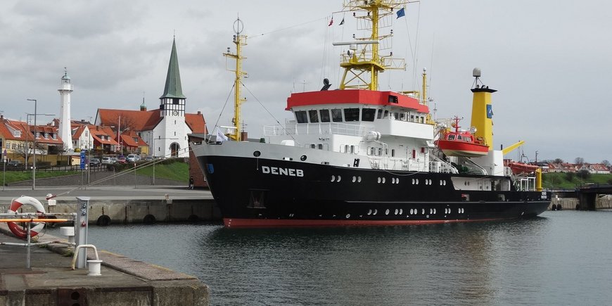 Dieses Foto zeigt das deutsche Vermessungsschiff „Deneb“ im Hafen der dänischen Stadt Bornholm. Der Betrachter schaut vom Hafenbecken aus schräg von vorn auf den Bug und die Backbordseite des Schiffes. Das Schiff ist etwa 100 Meter vom Betrachter entfernt. Es ist entlang seiner Steuerbordseite an der Hafenmauer festgemacht und man sieht etwa 1 Kilometer entfernt im Hintergrund die Kirche von Bornholm und daneben Häuser. Zwischen den Häusern steht ein Leuchtturm. Das Schiff ist ca. 52 Meter lang, 11 Meter breit und mit dem Mast mehr als 20 Meter hoch. Am Bug trägt es die Aufschrift „DENEB“.