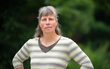 Portrait photo: A woman with long hair and fringes and a striped top stands in front of green trees.