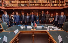 Group photo: 13 people stand in front of a wall of books, in the foreground are tables and the flags of Kazakhstan and Germany.