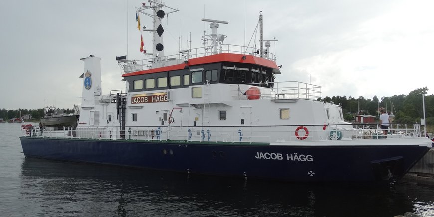 Dieses Foto zeigt das Schwedische Vermessungsschiff „Jacob Hägg“ in einem Hafen in der Nähe von Gävle. Es ist etwa 36 Meter lang und 7,5 Meter breit und mit dem Mast 15 Meter hoch. Das Schiff ist vom Betrachter etwa 50 Meter entfernt. Der Betrachter schaut vom Rand des Hafenbeckens aus schräg von vorn auf den Bug und die Steuerbordseite des Schiffes. Das Schiff ist entlang seiner Backbordseite an der Hafenmauer festgemacht. Am Bug und seitlich an der Brücke trägt es die Aufschrift „JACOB HÄGG“. Am Schornstein ist das Wappen der Schwedische Schifffahrtsbehörde mit Anker und Königskrone angebracht. Im Hintergrund sieht man den Wald der Küstenregion.