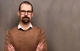 Sascha Brune, man with brown hair, glasses and beard in brown jumper with white collar, brown background