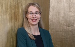 Portrait of Susanne Glaser: Woman with long blonde hair in a petrol-coloured jacket in front of a golden wall.