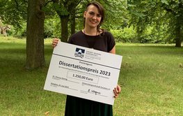 A person stands with a large cheque in his hand on a green lawn, trees in the background.