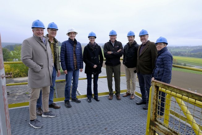 Eight people wearing hard hats stand on a platform of the drilling rig. Hilly landscape in the background.