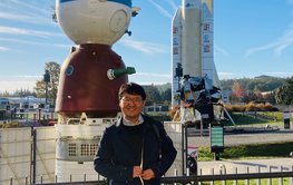 A young man with glasses stands smiling in a park in front of a Soyuz spacecraft and an Ariane space rocket. There are long white clouds and vapour trails in the blue sky.