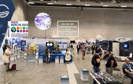 A trade fair stand in front of a grey wall. A large glowing globe hangs from the ceiling, people stand at tables below, others stroll through the hall.