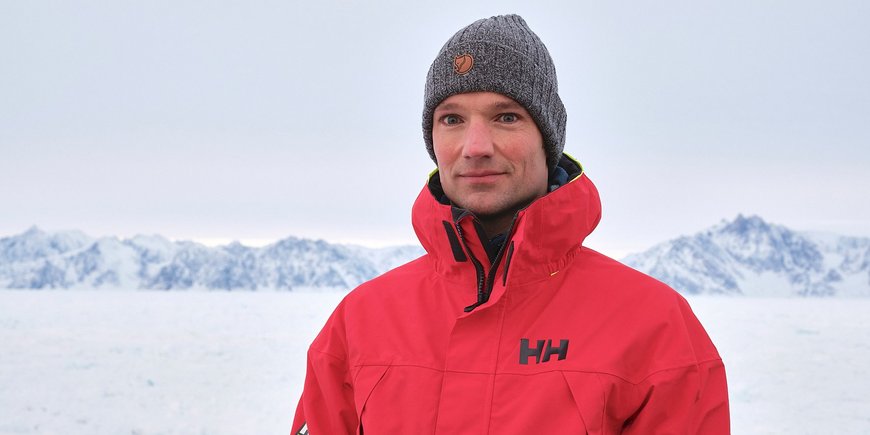 Tim Kalvelage, wearing a red functional jacket, sits in front of a snow-covered white background, with snow-capped mountains on the horizon and just a few rocks jutting out into the sky.