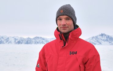 Tim Kalvelage szeht mit roter Funktionsjacke vor einem schneebedeckten weißen Hintergrund, am Horizont schneebedeckte Berge, aus denen nur ein paar Felsen in den Himmel ragen.