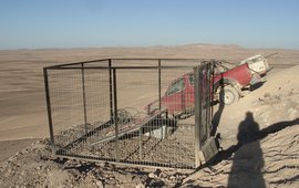 The photo shows a fenced-in solar installation in a dusty landscape. There is a car behind the fence