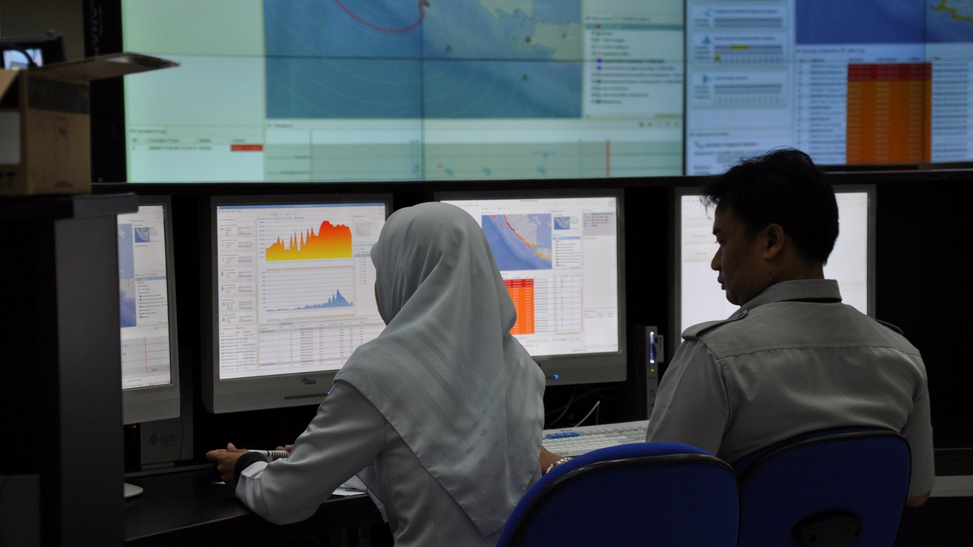 2 people sit in front of screens in the control room