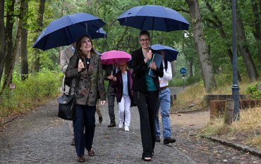 A group of people with umbrellas walk along a paved street through a park-like area.