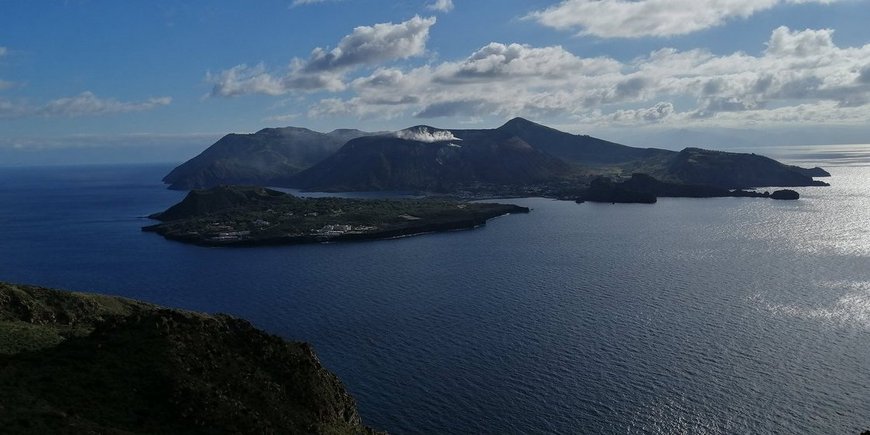 A dark grey volcanic island in the Mediterranean; blue sky with clouds. Vulcano island seen from the INGV Observatory in Lipari.