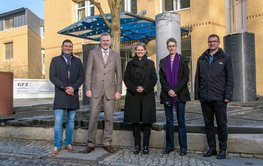 Group photo: Five people stand in front of stone pillars in front of the entrance to a yellow building.