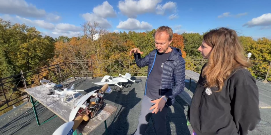 Screenshot from the live switch: A man and a woman stand on the roof of a building and explain a drone. An autumnal forest in the background.