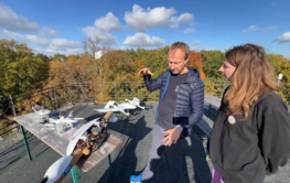 Screenshot from the live switch: A man and a woman stand on the roof of a building and explain a drone. An autumnal forest in the background.