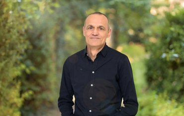 Portrait of a man in a black shirt and black trousers, his hands folded in front of his body. Green bushes and hedges in the background.
