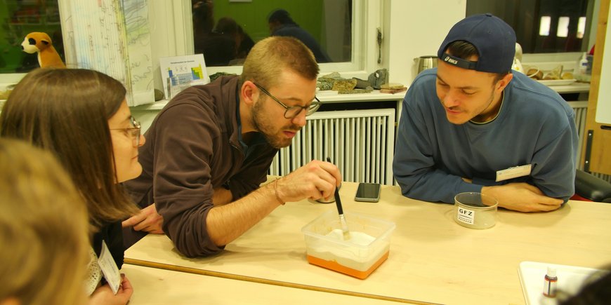 Um einen Tisch herum sitzen mehrere Menschen. Einer von ihnen verschiebt mit einem Pinsel Sand in einer kleinen Kiste, die auf dem Tisch steht.