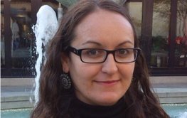 Portrait photo of a friendly smiling woman with long dark hair and dark glasses. A fountain in the background.