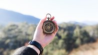 A hand holds an antique brass compass in front of a blurred mountain landscape. The hand is wearing a black fitness tracker on its wrist and a dark sleeve. The compass shows its detailed wind rose with clear markings against the blurred background of trees and mountains.